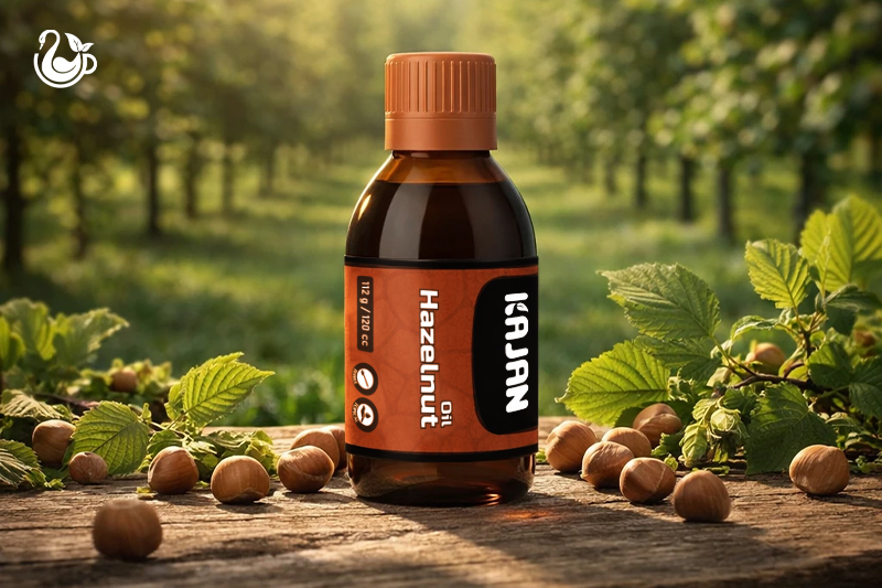 A bottle of Kajan Hazelnut oil with a red label and cap sits on a wooden surface. Near the bottle , hazelnuts can be seen.